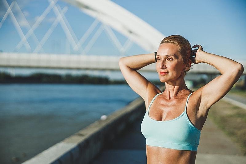 Woman stretching by a riverbank under bridge.